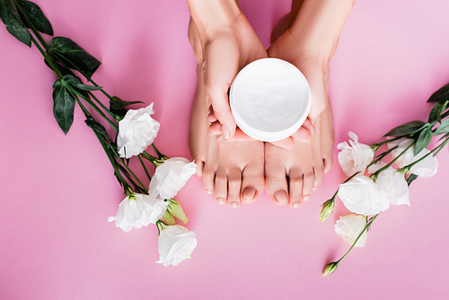 top view of female hands with cosmetic cream near feet and white eustoma flowers on pink background