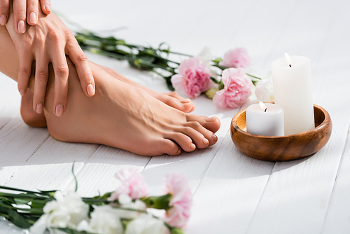cropped view of woman with groomed hands and feet near candles and flowers on white wooden surface, blurred foreground