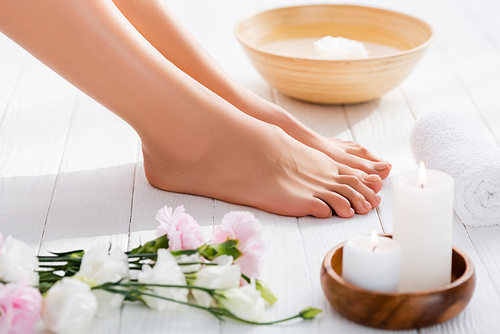 groomed female feet near eustoma flowers, candles and rolled towel on white wooden surface