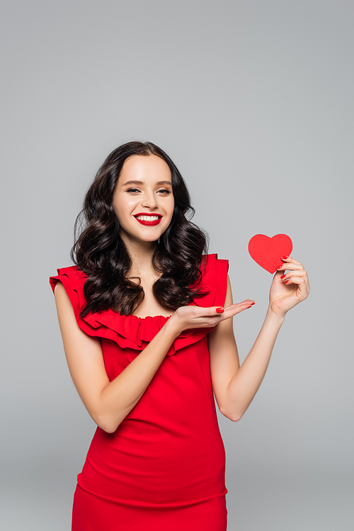 happy young woman pointing with hand at red paper heart isolated on grey