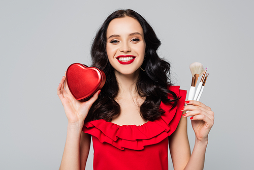 cheerful woman with red lips holding heart-shaped gift box and cosmetic brushes isolated on grey