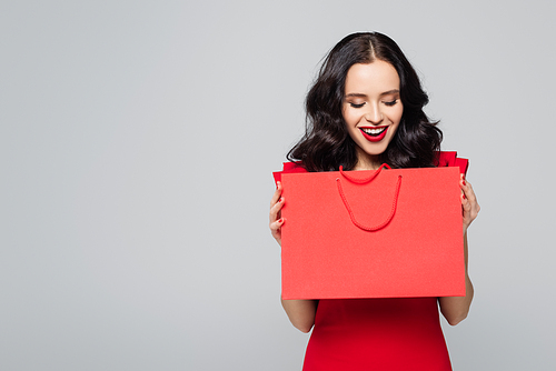 cheerful woman looking at purchase in shopping bag isolated on grey