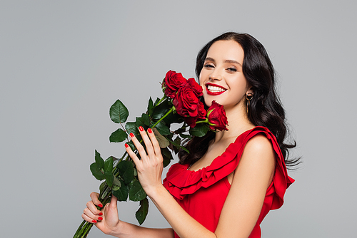cheerful young woman holding red roses and smiling isolated on grey