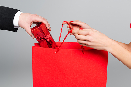 cropped view of man putting gift box in paper bag in hands of woman with red lips isolated on grey