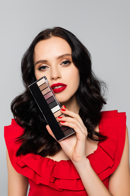 young brunette woman holding eye shadow palette isolated on grey