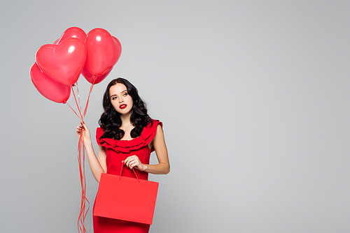 brunette woman holding red heart-shaped balloons and shopping bag isolated on grey