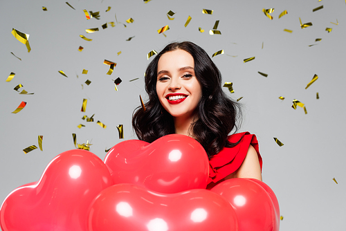 happy woman smiling near red heart-shaped balloons and falling confetti on grey
