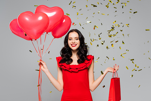 happy woman holding red heart-shaped balloons and shopping bag near falling confetti on grey