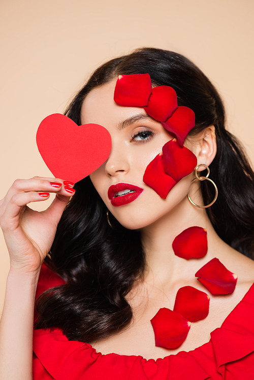 pretty young woman with petals of red rose on face covering eye with paper heart isolated on pink