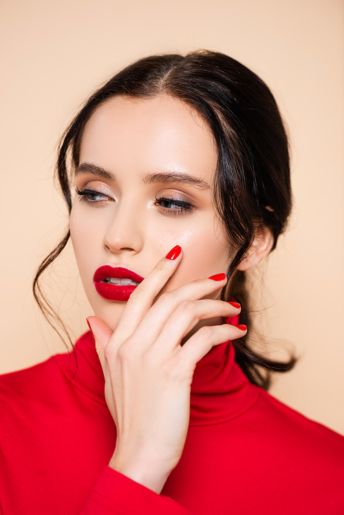 brunette young woman with red lips looking away while touching face isolated on pink