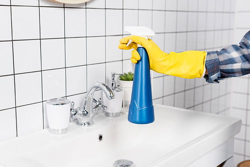 Cropped view of woman in rubber glove holding bottle of detergent near sink and tile in bathroom