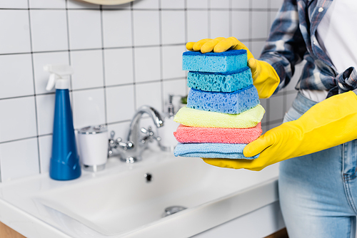 Cropped view of woman in rubber gloves holding colorful sponges in bathroom