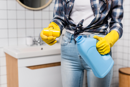 Cropped view of bottle of cleaner and cap in hands of woman in rubber gloves on blurred background in bathroom