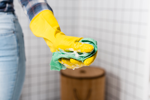 Cropped view of rag with soap in hand of woman in rubber glove on blurred background in bathroom
