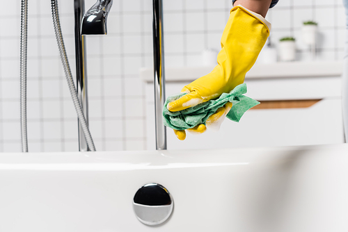 Cropped view of woman in rubber glove holding rag with soap near faucet and bathtub