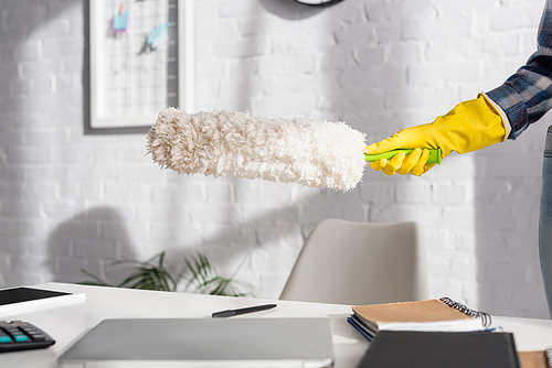 Cropped view of woman in rubber glove holding dust brush near devices and notebook on blurred foreground on table