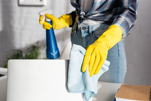 Cropped view of woman in rubber gloves cleaning laptop with rag and holding detergent on blurred background