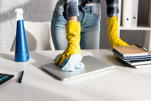 Cropped view of woman in rubber gloves cleaning laptop near detergent and notebooks on blurred background on table
