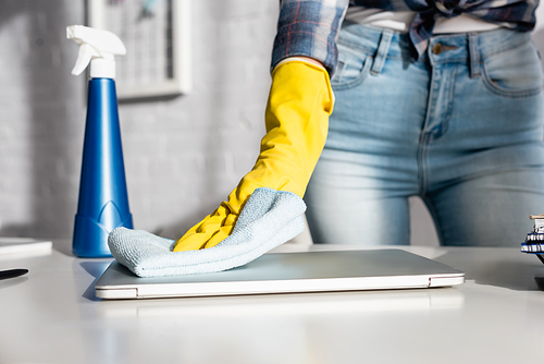 Cropped view of woman in rubber glove cleaning laptop near bottle of detergent on blurred background at home