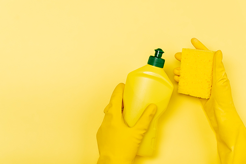 Top view of hands in rubber gloves holding bottle of dishwashing liquid and sponge on yellow background