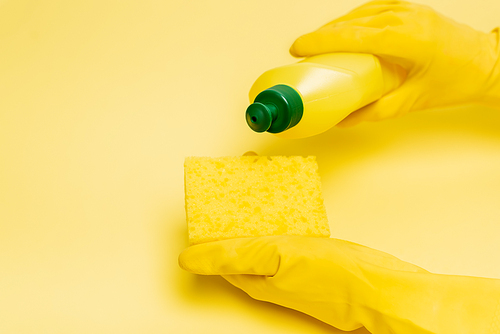 Cropped view of hands in rubber gloves pouring dishwashing liquid on sponge on yellow background