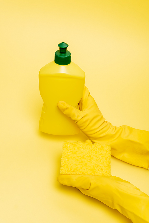 High angle view of hands in rubber gloves holding sponge and dishwashing liquid on yellow background