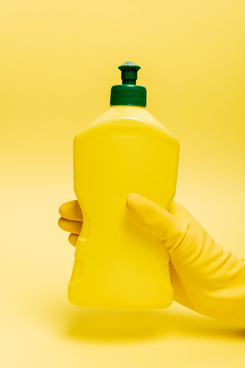 Cropped view of hand in rubber glove holding bottle of dishwashing liquid on yellow background