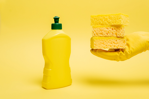 Cropped view of person in rubber glove holding sponges near dishwashing liquid on yellow background