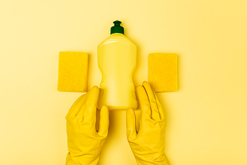 Top view of person in rubber gloves holding dishwashing liquid near sponges on yellow background