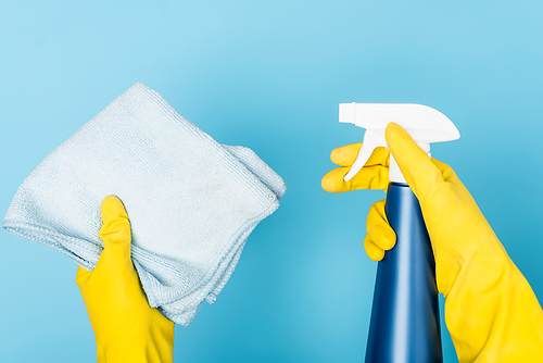 Cropped view of hands in rubber gloves holding detergent with sprayer and rag on blue background