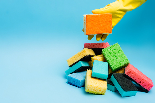 Cropped view of person in yellow rubber glove holding sponge near sponges on blue background