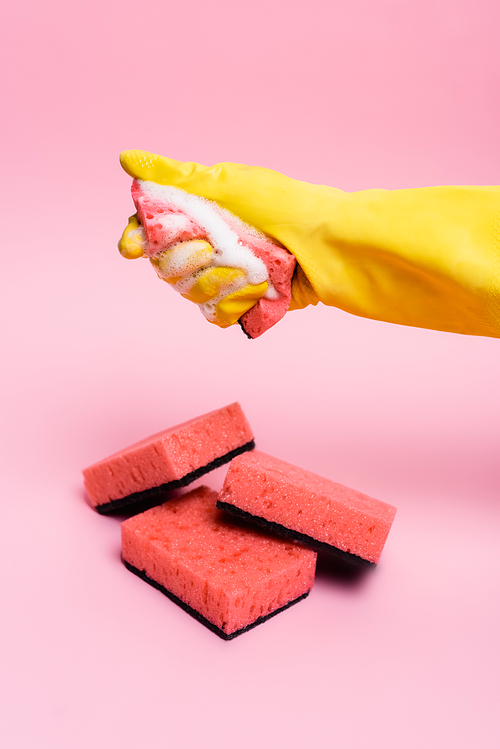 Hand in rubber glove holding sponge with soapsuds near sponges on pink background