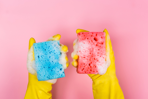 Top view of hands in rubber gloves holding sponges with soap foam on pink background