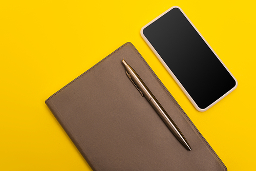 top view of smartphone with blank screen near copy book and golden pen isolated on yellow