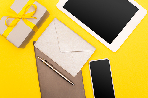 top view of gadgets with blank screen, copy book with golden pen and envelope near present isolated on yellow