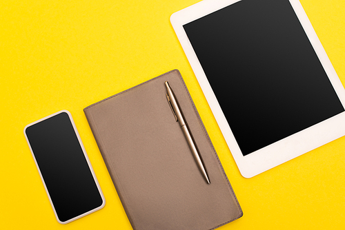 top view of gadgets with blank screen near copy book with golden pen isolated on yellow