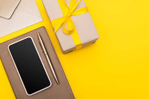 top view of smartphone with blank screen on copy book with pen near envelope and gift box isolated on yellow