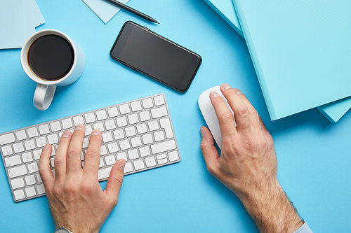 Cropped view of man using computer keyboard and computer mouse at workplace with papers, smartphone and cup of coffee on blue background