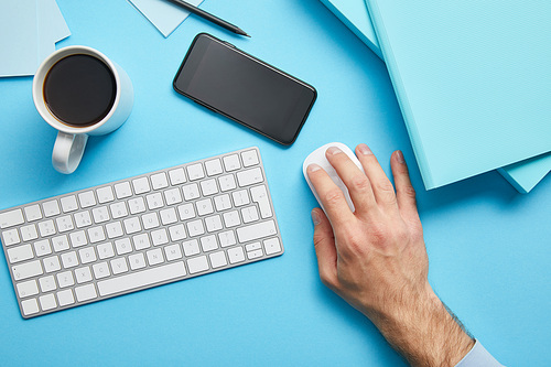 Cropped view of man using computer mouse at workplace with papers and cup of coffee on blue background
