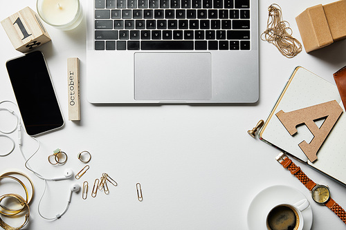 top view of laptop, smartphone, jewelry, case, earphones, candle, flowers, office supplies and coffee on white surface