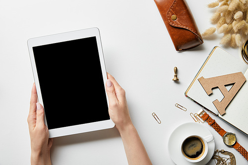 cropped view of digital tablet in woman hands on white surface with stationery, jewelry, notepad, case and coffee