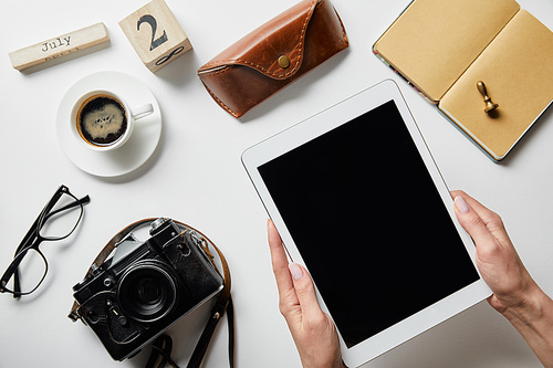 cropped view of woman holding digital tablet near photo camera, coffee, case, notepad, block with numbers and letters on white surface