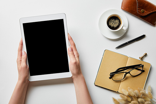cropped view of woman holding digital tablet in hands near notepad, coffee, glasses and case on white surface