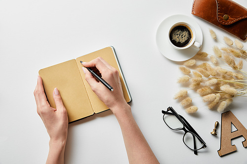 cropped view of woman writing in notepad near coffee, glasses and case on white surface