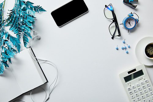 top view of smartphone near notepad, coffee, stationery, earphones, alarm clock, calculator, glasses and blue branch on white surface