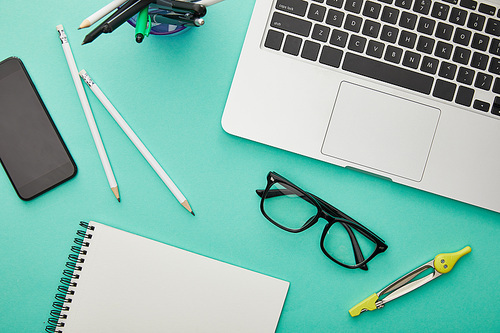 top view of gadgets near glasses, stationery and notebook isolated on turquoise