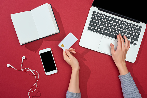 top view of credit card on red background with smartphone, laptop, earphones, coffee, notebook and plant