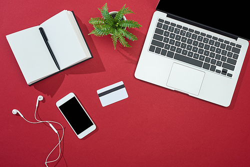 top view of credit card, smartphone, laptop, earphones, pen, notebook and plant on red background