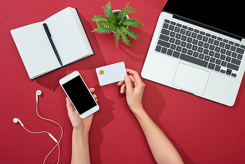 cropped view of woman holding credit card and smartphone near laptop, earphones, pen, notebook and plant on red background