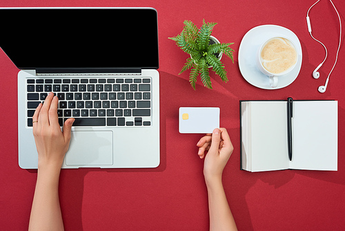 cropped view of woman holding credit card and using laptop near earphones, coffee, notebook with pen and plant on red background
